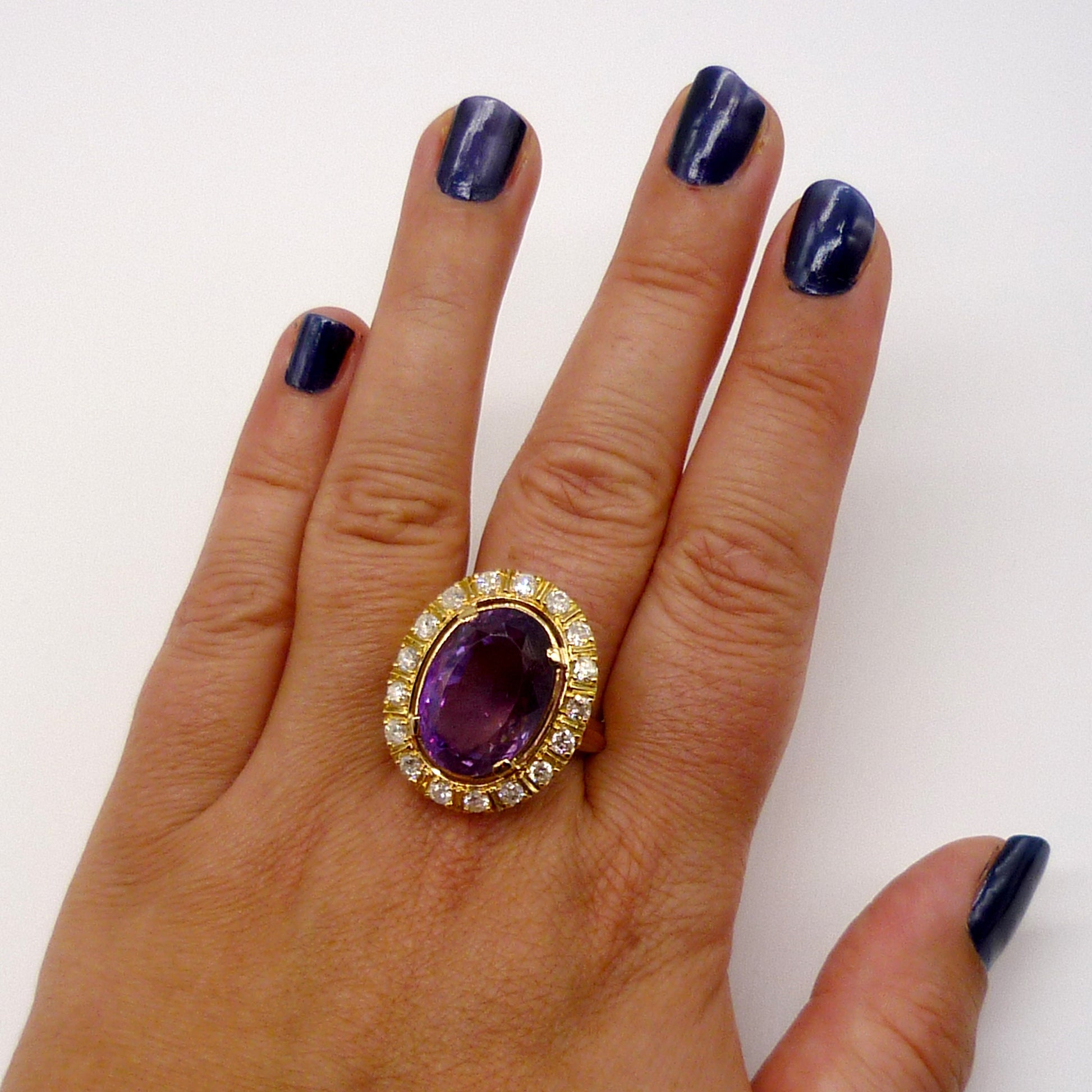 A very large purple amethyst ring with diamond and yellow gold surround on the middle finger of a woman's hand, viewed from above. The finger nails are painted dark blue.