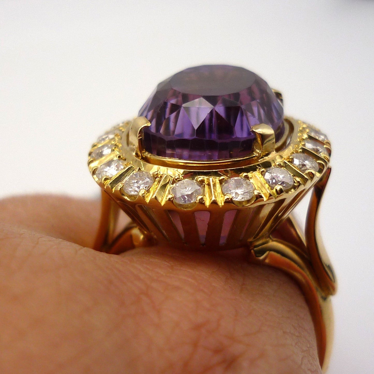 A huge purple amethyst ring with diamond and yellow gold surround, worn on a finger. The view is taken from the side and the ring sits high up on an open gallery